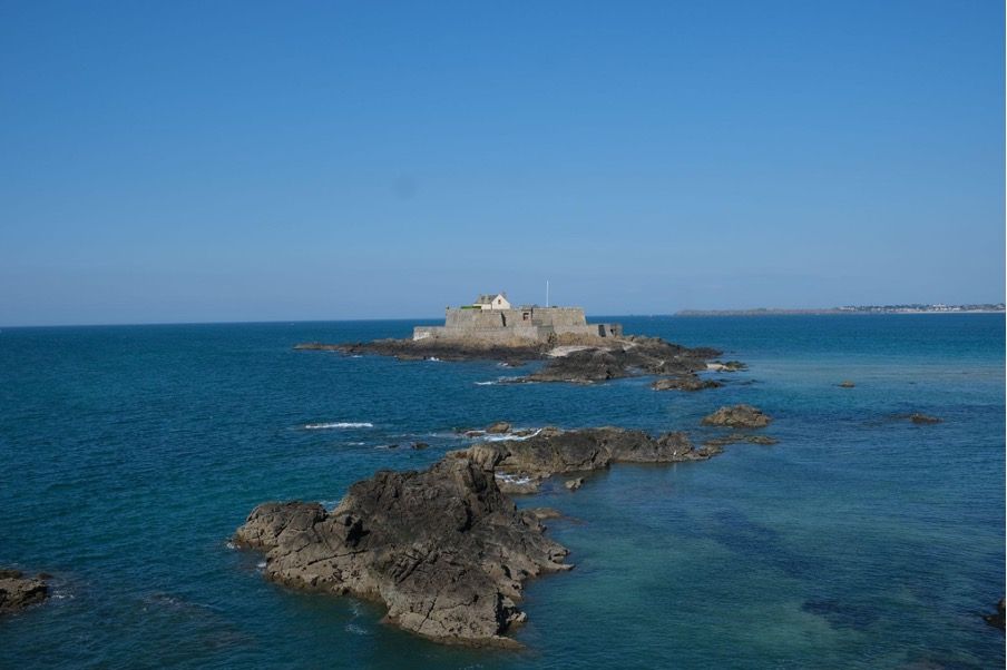 Photo of rocks in the sea, with a tiny island in which there’s a house, surrounded by fortification walls.