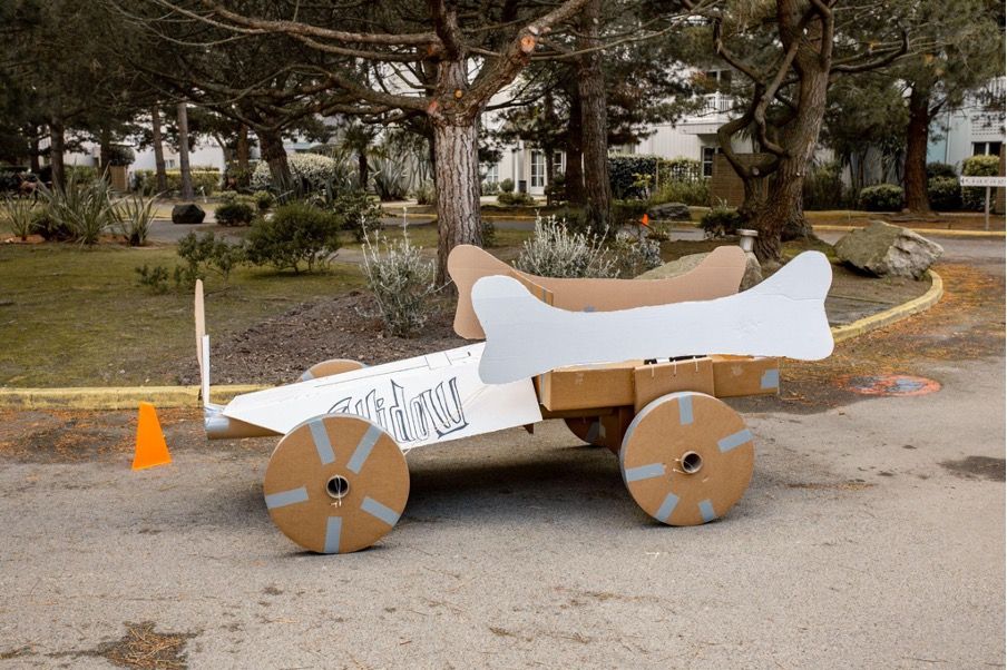 A white kart made all in cardboard on the road in front of trees. The kart sides are decorated with bone shapes.