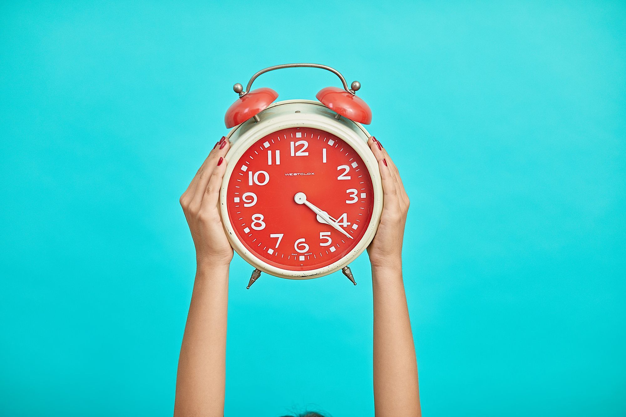 A pair of hands holding up a large red alarm clock with a white frame, set against a bright turquoise background. The clock displays 4:22 time. The person's nails are painted in a vibrant red to match the clock's face.